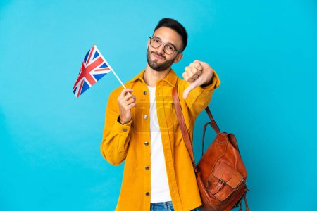 Foto de Joven hombre caucásico sosteniendo una bandera del Reino Unido aislado sobre fondo amarillo mostrando el pulgar hacia abajo con expresión negativa - Imagen libre de derechos