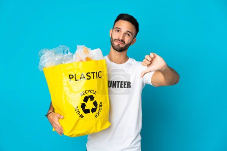 Photo for Young caucasian man holding a bag full of plastic bottles to recycle isolated on blue background showing thumb down with negative expression - Royalty Free Image