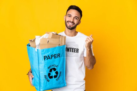 Foto de Joven hombre caucásico sosteniendo una bolsa de reciclaje llena de papel para reciclar aislado sobre fondo blanco haciendo gesto de dinero - Imagen libre de derechos