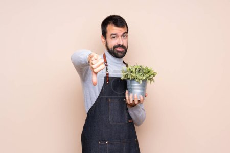 Téléchargez les photos : Homme tenant une plante sur fond isolé montrant le pouce baissé avec une expression négative - en image libre de droit