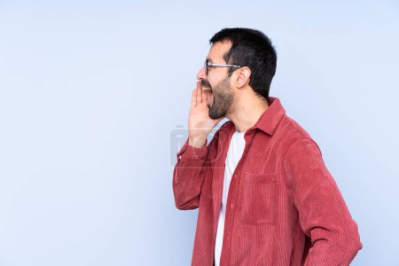 Foto de Joven hombre caucásico con chaqueta de pana sobre fondo azul gritando con la boca abierta hacia el lateral - Imagen libre de derechos