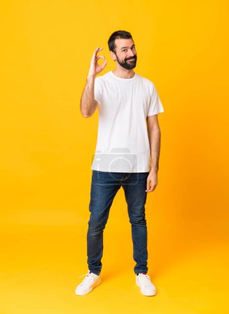 Téléchargez les photos : Prise de vue pleine longueur de l'homme avec barbe sur fond jaune isolé montrant ok signe avec les doigts - en image libre de droit