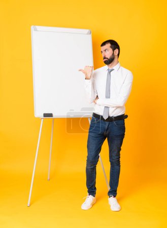 Téléchargez les photos : Plan complet de l'homme d'affaires donnant une présentation sur tableau blanc sur fond jaune isolé malheureux et pointant vers le côté - en image libre de droit