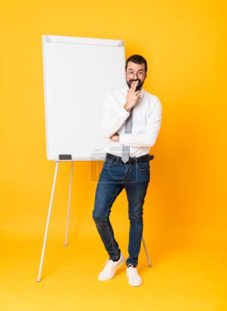Téléchargez les photos : Plan complet de l'homme d'affaires donnant une présentation sur tableau blanc sur fond jaune isolé avec des lunettes et surpris - en image libre de droit