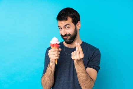 Téléchargez les photos : Jeune homme avec une crème glacée cornet sur fond bleu isolé faisant geste d'argent - en image libre de droit
