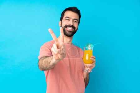 Téléchargez les photos : Jeune homme tenant un cocktail sur fond bleu isolé souriant et montrant signe de victoire - en image libre de droit