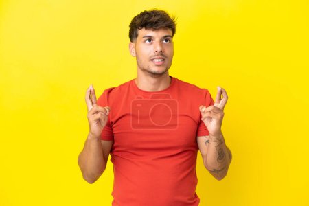 Téléchargez les photos : Jeune homme beau caucasien isolé sur fond jaune avec les doigts croisés et souhaitant le meilleur - en image libre de droit