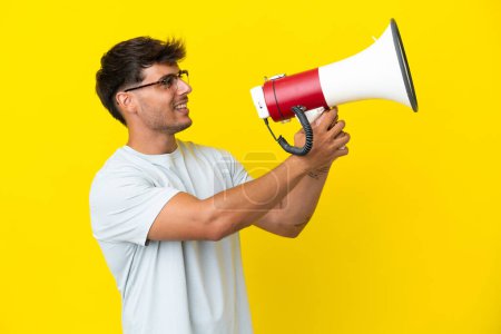 Téléchargez les photos : Jeune homme beau caucasien isolé sur fond jaune criant à travers un mégaphone pour annoncer quelque chose - en image libre de droit