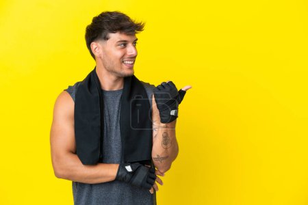 Foto de Joven deportista caucásico con toalla aislada sobre fondo amarillo apuntando hacia un lado para presentar un producto - Imagen libre de derechos