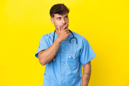 Téléchargez les photos : Jeune chirurgien médecin homme isolé sur fond jaune ayant des doutes et avec expression confuse visage - en image libre de droit