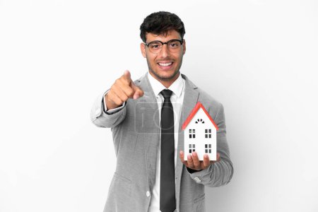 Téléchargez les photos : Homme d'affaires tenant une maison isolée sur fond bleu pointant devant avec expression heureuse - en image libre de droit