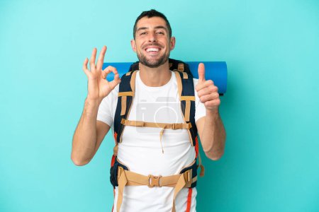 Photo for Young mountaineer caucasian man with a big backpack isolated on blue background showing ok sign and thumb up gesture - Royalty Free Image