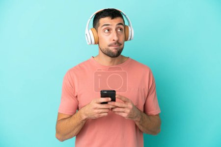 Téléchargez les photos : Jeune homme caucasien beau isolé sur fond bleu écoutant de la musique avec un mobile et pensant - en image libre de droit
