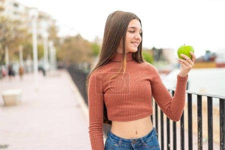 Téléchargez les photos : Adolescente avec une pomme à l'extérieur avec une expression heureuse - en image libre de droit