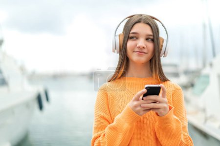 Téléchargez les photos : Adolescente à l'extérieur écoutant de la musique avec un mobile et pensant - en image libre de droit