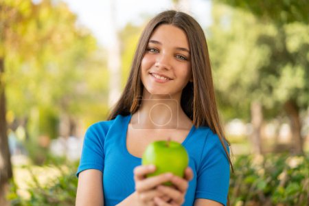 Téléchargez les photos : Adolescente à l'extérieur tenant une pomme avec une expression heureuse - en image libre de droit