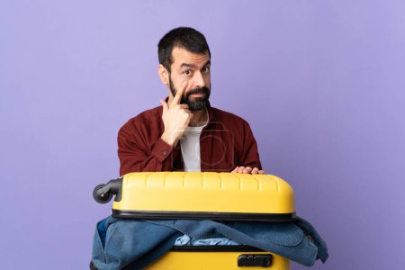 Foto de Viajero caucásico hombre con una maleta llena de ropa sobre aislado púrpura fondo mostrando algo - Imagen libre de derechos