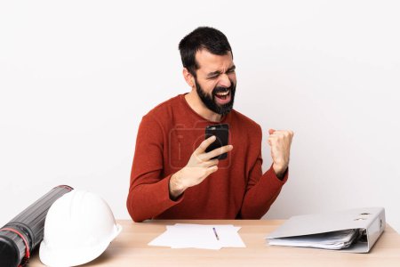 Photo for Caucasian architect man with beard in a table with phone in victory position. - Royalty Free Image