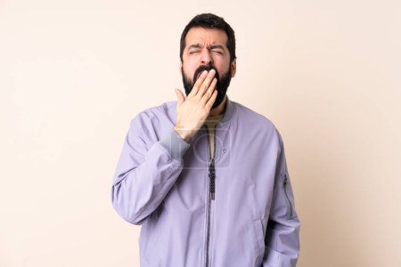 Téléchargez les photos : Homme caucasien avec barbe portant une veste sur fond isolé bâillant et couvrant la bouche grande ouverte avec la main - en image libre de droit