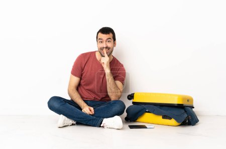 Foto de Hombre guapo caucásico con una maleta llena de ropa sentada en el suelo en interiores mostrando un gesto de silencio poniendo el dedo en la boca - Imagen libre de derechos