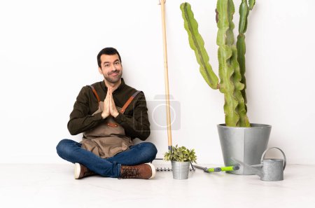 Photo for Gardener man sitting on the floor at indoors keeps palm together. Person asks for something - Royalty Free Image