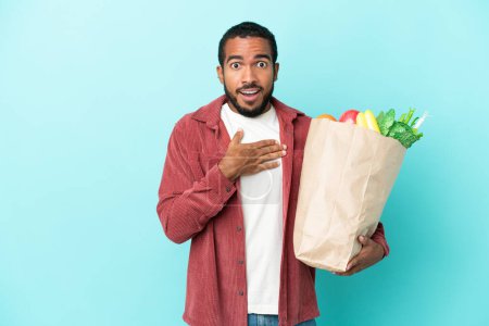 Foto de Joven latino sosteniendo una bolsa de la compra de comestibles aislado sobre fondo azul sorprendido y sorprendido mientras mira a la derecha - Imagen libre de derechos