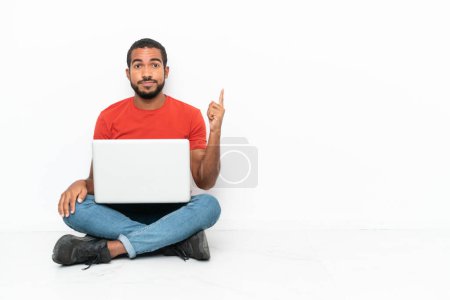 Foto de Joven ecuatoriano con un portátil sentado en el suelo aislado sobre fondo blanco señalando con el dedo índice una gran idea - Imagen libre de derechos