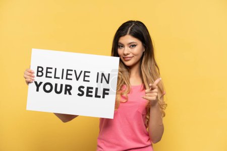 Téléchargez les photos : Jeune fille russe isolée sur fond jaune tenant une pancarte avec le texte Believe In Your Self et pointant vers l'avant - en image libre de droit