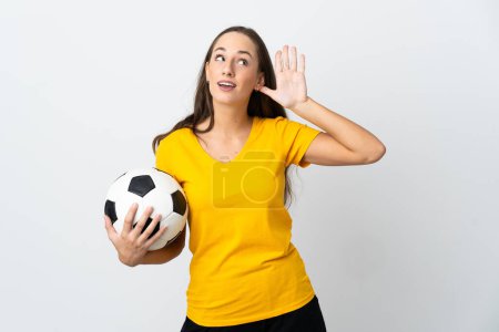 Téléchargez les photos : Jeune footballeuse Femme sur fond blanc isolé écoutant quelque chose en mettant la main sur l'oreille - en image libre de droit