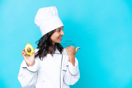 Foto de Joven chef hispana sosteniendo aguacate aislado sobre fondo azul apuntando hacia un lado para presentar un producto - Imagen libre de derechos