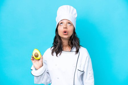 Photo for Young hispanic chef woman holding avocado isolated on blue background looking up and with surprised expression - Royalty Free Image