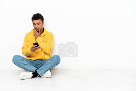 Téléchargez les photos : Jeune homme afro-américain assis sur le sol isolé sur fond blanc penser et envoyer un message - en image libre de droit