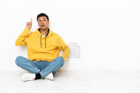 Foto de Joven hombre afroamericano sentado en el suelo aislado sobre fondo blanco con la intención de realizar la solución mientras levanta un dedo - Imagen libre de derechos