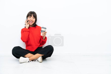Téléchargez les photos : Jeune femme métisse assise sur le sol isolée sur fond blanc tenant du café à emporter et un mobile - en image libre de droit