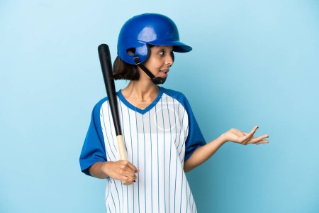 Foto de Béisbol mujer jugador de raza mixta con casco y murciélago aislado sobre fondo azul con expresión sorpresa mientras mira al lado - Imagen libre de derechos