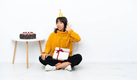 Téléchargez les photos : Jeune femme métisse tenant un cadeau assis sur le sol isolé sur fond blanc écoutant quelque chose en mettant la main sur l'oreille - en image libre de droit