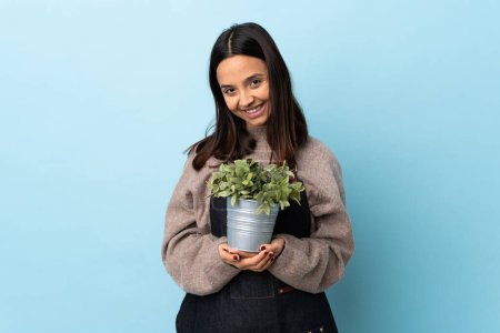Photo for Young brunette mixed race woman holding a plant over isolated blue background keeps palm together. Person asks for something. - Royalty Free Image