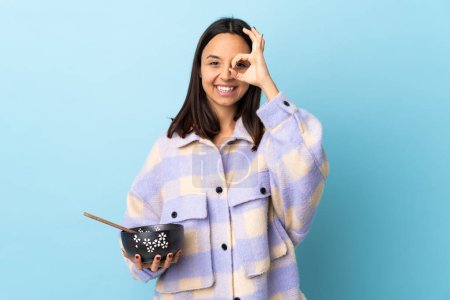 Téléchargez les photos : Jeune brune mixte femme tenant un bol plein de nouilles sur fond bleu isolé montrant ok signe avec les doigts. - en image libre de droit