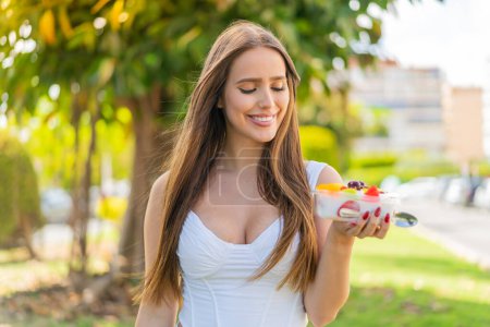 Foto de Mujer joven sosteniendo un tazón de fruta al aire libre con expresión feliz - Imagen libre de derechos