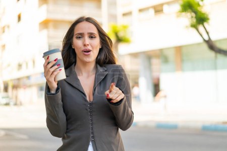 Téléchargez les photos : Jeune femme tenant un café à emporter à l'extérieur surpris et pointant devant - en image libre de droit