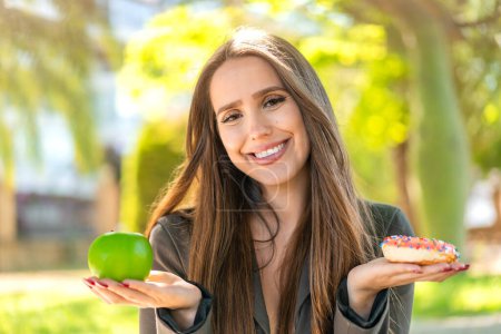 Téléchargez les photos : Jeune femme à l'extérieur tenant pomme et beignet avec une expression heureuse - en image libre de droit