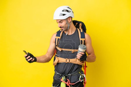 Foto de Joven escalador caucásico aislado sobre fondo amarillo sosteniendo café para llevar y un móvil - Imagen libre de derechos
