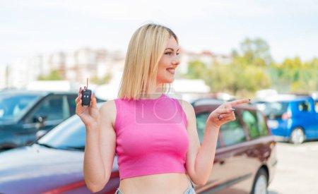 Foto de Joven mujer rubia bonita sosteniendo las llaves del coche al aire libre apuntando a un lado para presentar un producto - Imagen libre de derechos