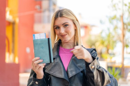 Téléchargez les photos : Jeune jolie femme blonde tenant un passeport à l'extérieur pointe du doigt vers vous avec une expression confiante - en image libre de droit