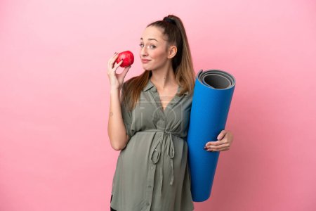 Téléchargez les photos : Jeune femme isolée sur fond rose enceinte tenant une pomme et allant à des cours de yoga - en image libre de droit