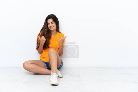 Photo for Young caucasian woman sitting on the floor isolated on white background with thumbs up gesture and smiling - Royalty Free Image