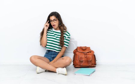 Téléchargez les photos : Jeune étudiante assise sur le sol isolé sur fond blanc avec des lunettes et surprise - en image libre de droit