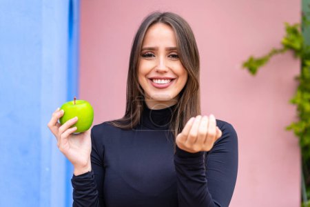 Téléchargez les photos : Jeune jolie femme avec une pomme à l'extérieur invitant à venir avec la main. Heureux que tu sois venu - en image libre de droit