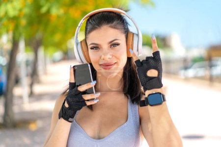 Téléchargez les photos : Jeune jolie femme sportive à l'extérieur écoutant de la musique avec un geste rock mobile - en image libre de droit