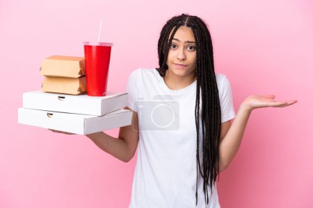 Téléchargez les photos : Adolescente avec des tresses tenant des pizzas et des hamburgers sur fond rose isolé ayant des doutes tout en levant la main - en image libre de droit